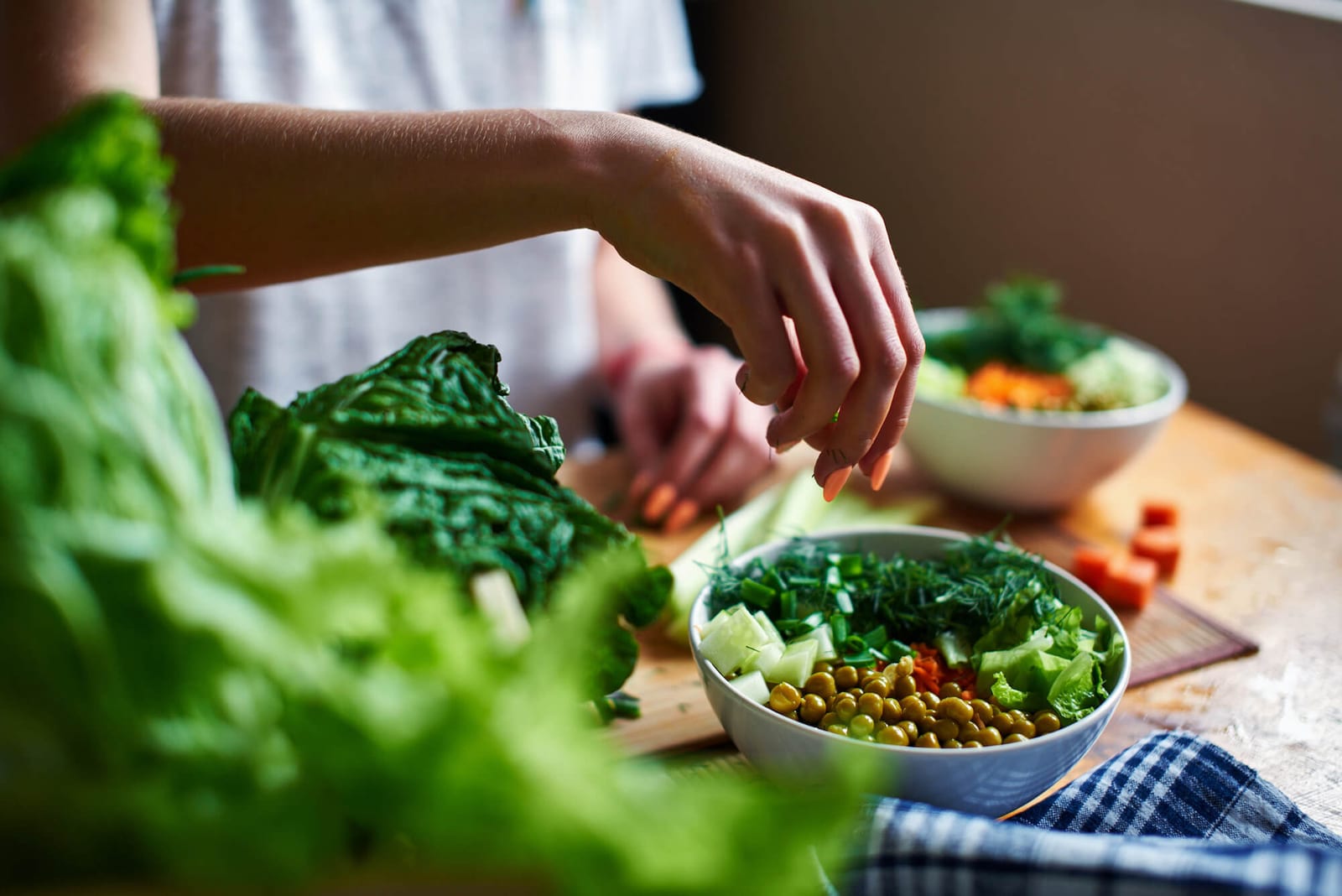 Female hand pours green onions in a bowl with green peas, cucumbers, carrots, lettuce and dill standing on a table as an example of how a dietitian can help you meet your health goals