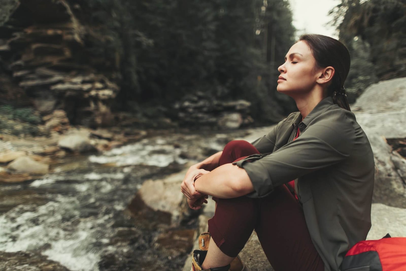 Peaceful young beautiful woman sitting on the stone near mountain river while resting after a long walk