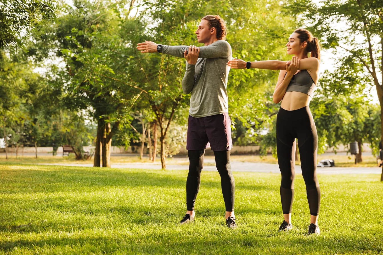Photo of fitness sportsman and sportswoman 20s in tracksuits warming up together and stretching arms in green park during sunny summer day
